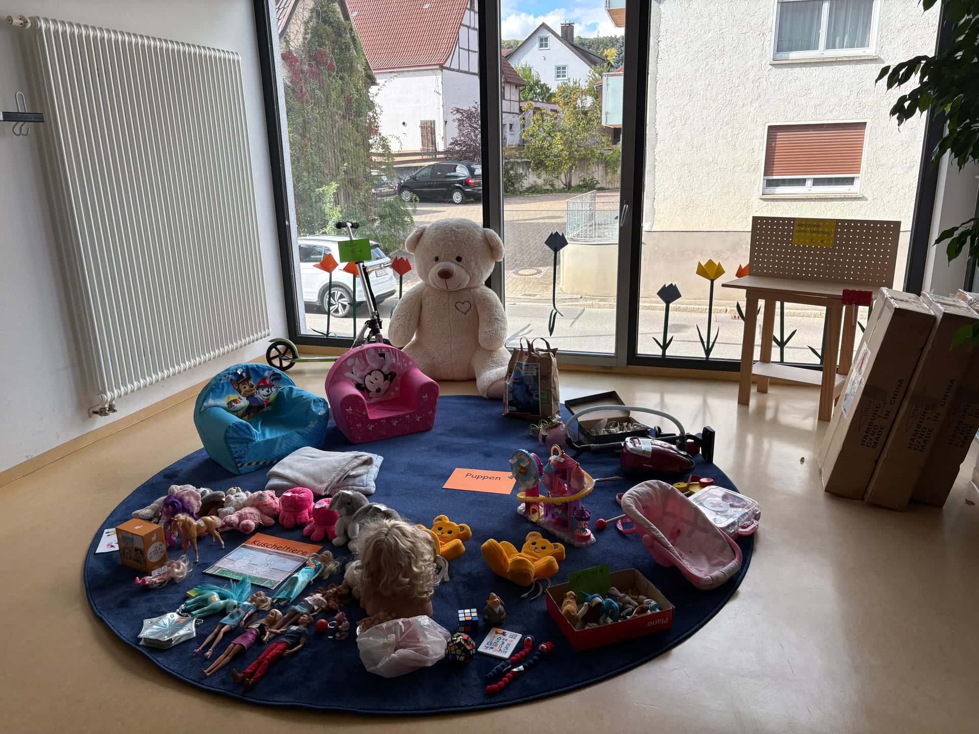 Flohmarkt 2026 - Puppen, Kuscheltiere und Spielsachen auf einem runden Teppich vor der Fensterfront, dahinter ein großer Teddybär