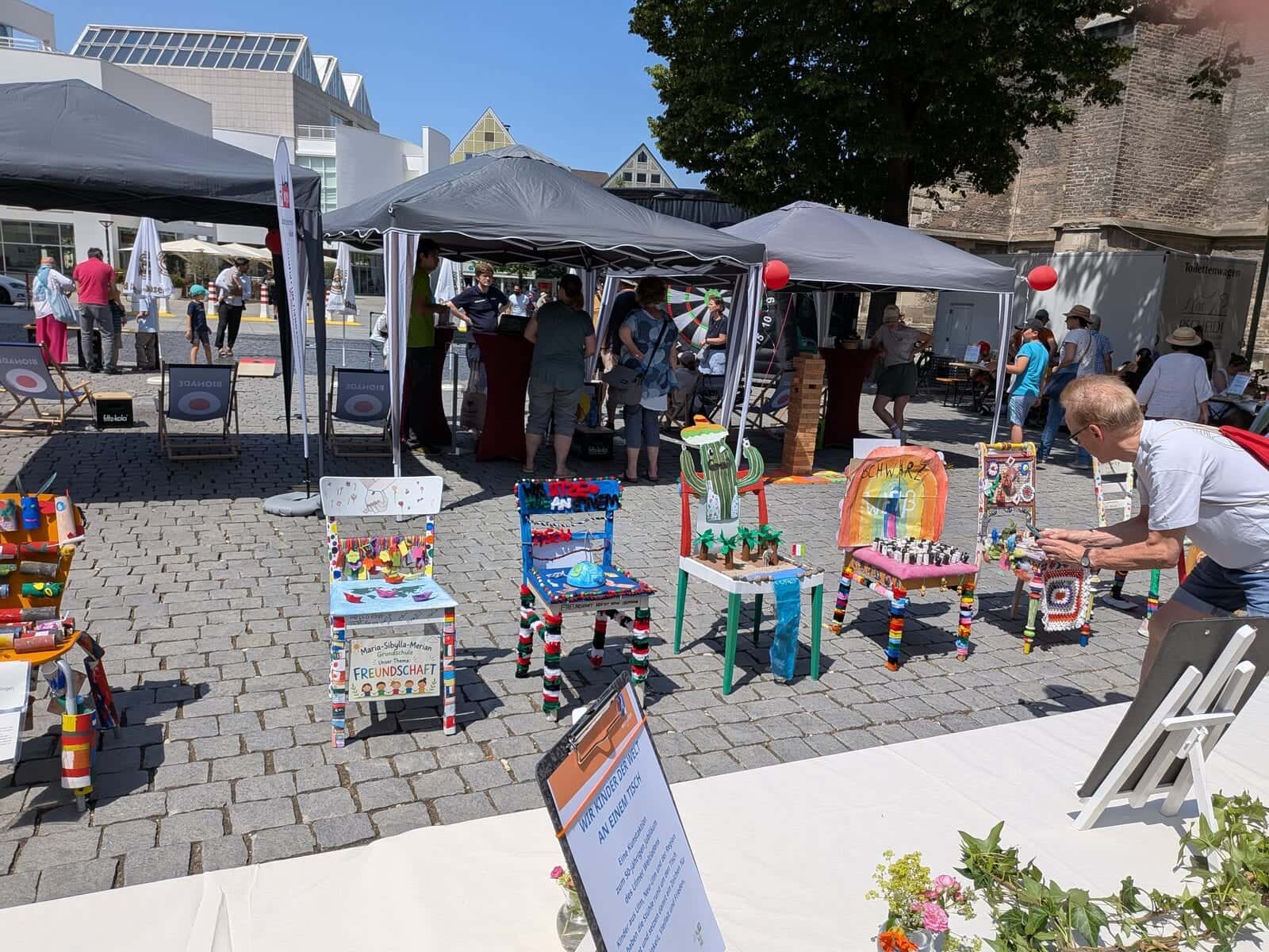 Wir sitzen alle an einem Tisch 2026 - Reihe bunt bemalter Stühle bei der Kunstaktion auf dem Münsterplatz unter Pavillonzelten