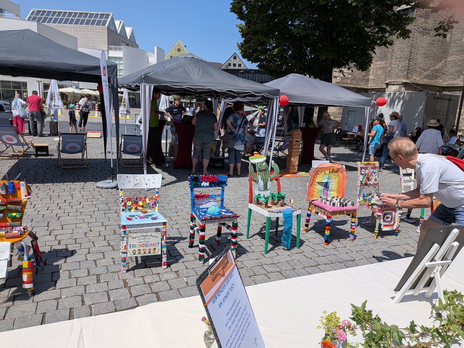 Wir sitzen alle an einem Tisch 2026 - Reihe bunt bemalter Stühle bei der Kunstaktion auf dem Münsterplatz unter Pavillonzelten